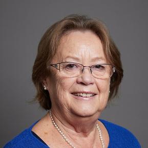 Portrait photo of Baroness Bakewell in front of a grey background. Baroness Bakewell is a white woman with short brown hair. She wears glasses and a blue cardigan.